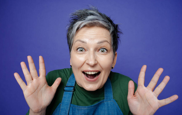 Close-up portrait of middle age grey haired pretty surprised woman with opened mouth standing with open palms isolated on blue background. Portrait of happy exited aged woman with with short haircut - Zdjęcie, obraz