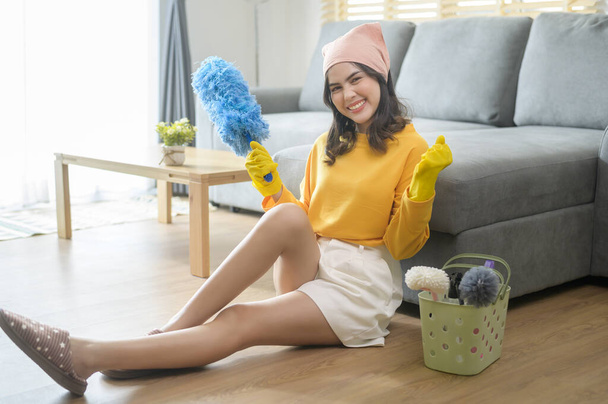 A Young happy woman wearing yellow gloves  and holding a basket of cleaning supplies in living room. - Фото, изображение