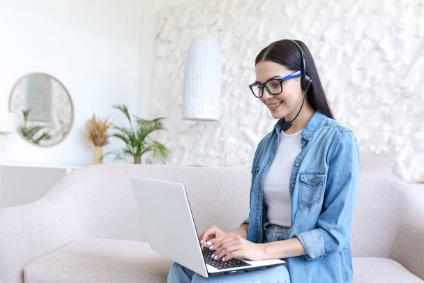 Young beautiful woman working at home sitting on sofa, using headset and laptop for video call, tech support of online store, and customer service worker - Foto, immagini