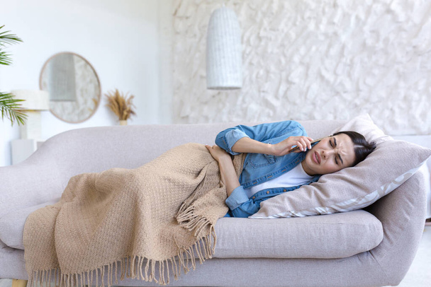 Upset sick woman alone at home, lying on the sofa under a blanket, calling the doctor on the phone - Φωτογραφία, εικόνα