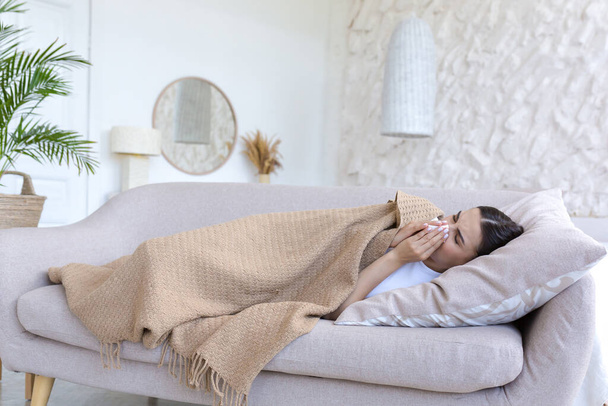 Uma mulher doente está deitada debaixo de um cobertor no sofá sozinha em casa, tem um resfriado, está espirrando - Foto, Imagem