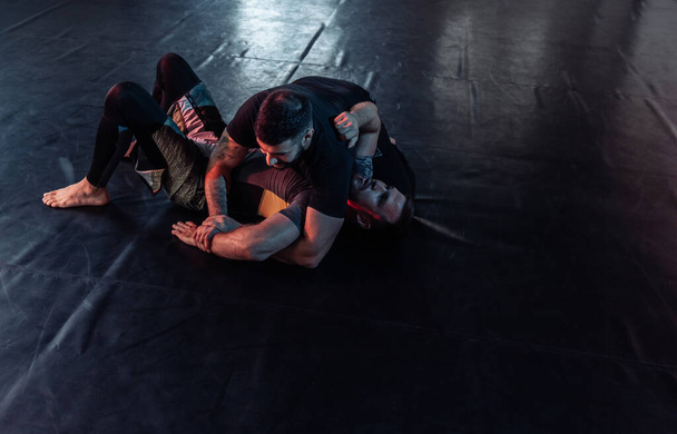 Twee atletische mannelijke vechters trainen, oefenen een pijnlijke greep in een sporthal. DArk neon licht - Foto, afbeelding