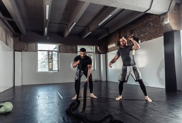 Функциональное обучение. Два спортсмена-мужчины тренируются с гирями и боевыми канатами в спортзале - Фото, изображение