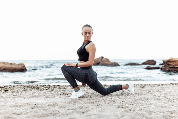 Красивая молодая женщина в спортивной одежде, практикующая растягивание ног в одиночестве на диком пляже. Понятие здорового образа жизни - Фото, изображение