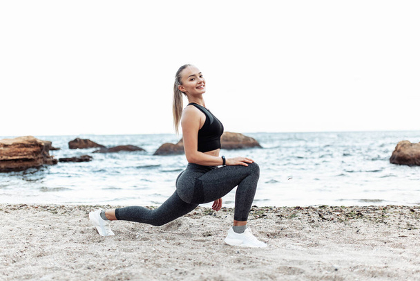 Beautiful young fit woman in sportswear practicing stretching legs alone on wild beach. Healthy lifestyle concept - Zdjęcie, obraz