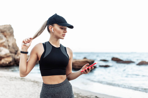 Fit woman in sportswear and a cap uses a smartphone on a wild beach - Foto, Imagem
