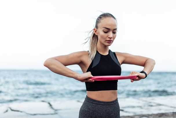 Young beautiful fit woman in sportwear training with a fitness elastic band (expander) on a urban beach - Fotografie, Obrázek