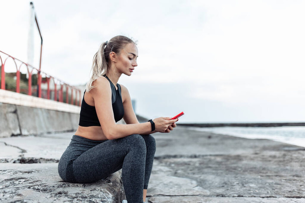 Fit woman in sportswear using smartphone while sitting on urban beach - Fotografie, Obrázek