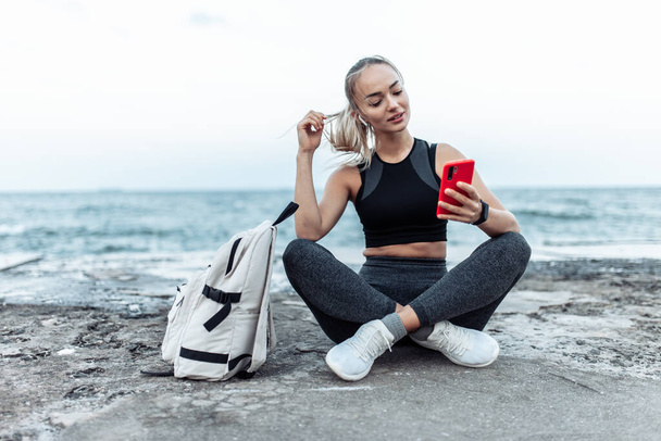 Happy Fit woman in sportswear using smartphone and listen music in headphones while sitting on urban beach - Foto, afbeelding