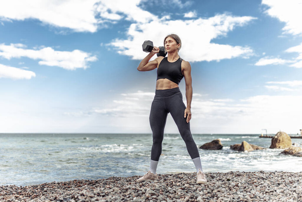 Strong athletic woman exercising with heavy dumbbell on the beach during the day with blue sky and clouds. Functional outdoor training - Foto, afbeelding