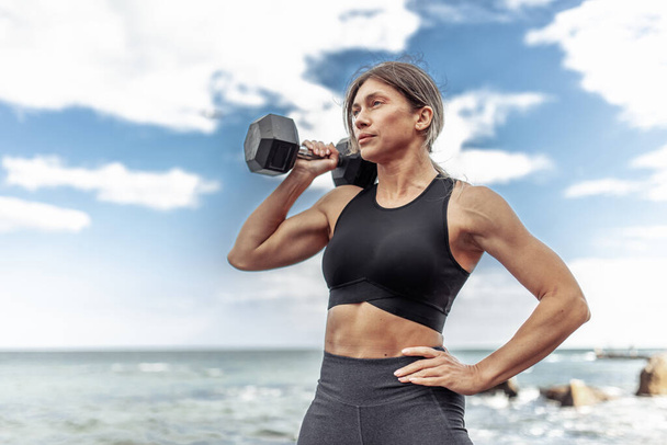 Strong athletic woman exercising with heavy dumbbell on the beach during the day with blue sky and clouds. Functional outdoor training - 写真・画像