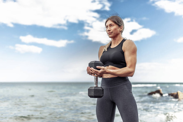 Strong athletic woman exercising with heavy dumbbell on the beach during the day with blue sky and clouds. Functional outdoor training - Foto, immagini