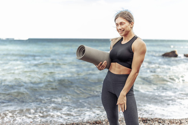 Happy Muscular woman athlete in sportswear posing with mat on the beach - Foto, Bild