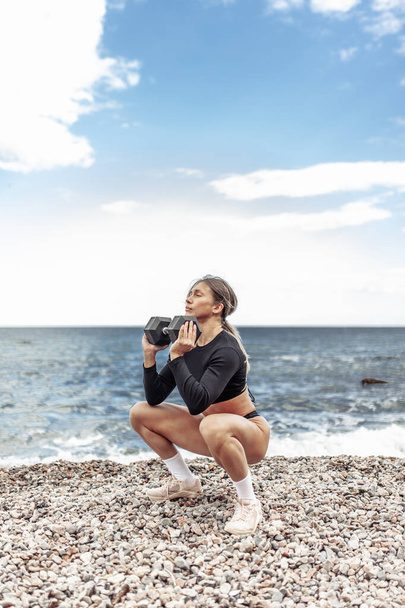 Strong athletic woman exercising with heavy dumbbell on the beach during the day with blue sky and clouds. Functional outdoor training - Foto, Imagen