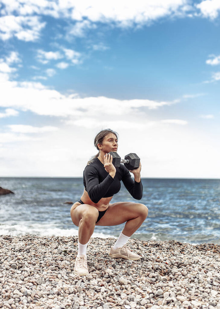 Strong athletic woman exercising with heavy dumbbell on the beach during the day with blue sky and clouds. Functional outdoor training - Fotó, kép