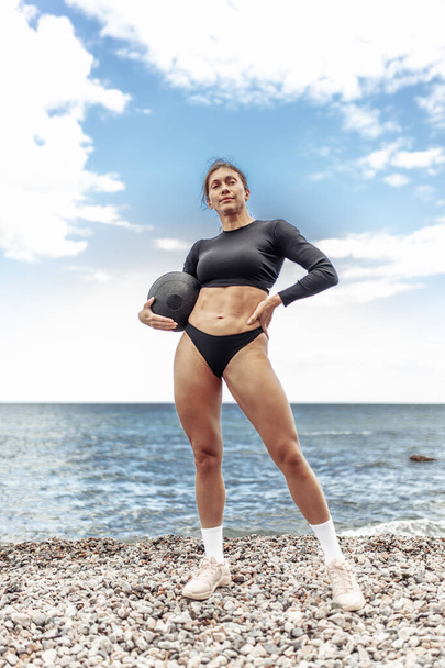 Μυώδης αθλήτρια στα αθλητικά που ποζάρει με ιατρικές μπάλες στην παραλία - Φωτογραφία, εικόνα
