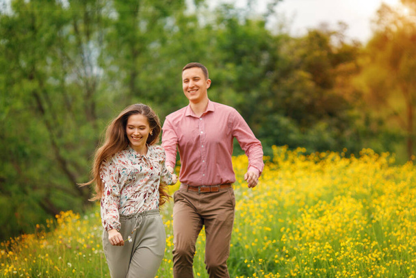  Heureux couple amoureux courent ensemble et se tenant la main, deux personnes profitant d'une promenade à travers la terre herbeuse. La jeune femme rit et ses cheveux flottent - Photo, image