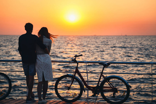 Couple of young hipster lovers hugging at the beach and watching the sunrise at wooden deck summer time. - Foto, Imagen