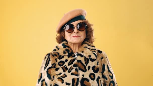 Stijlvolle senior vrouw in een zonnebril met een luipaard bontjas en een baret terwijl ze glimlacht over een gele achtergrond. - Video