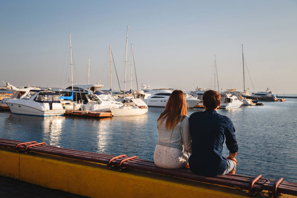 Cute young beautiful couple sitting and hugging together at pier at port, hipsters. Back view. - Fotoğraf, Görsel