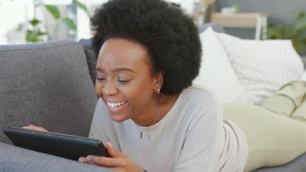 Laughing afro woman using digital tablet to stream funny movies online on subscription channel. Smiling, happy woman lying on home living room sofa, relaxing and using technology for online shopping. - Πλάνα, βίντεο