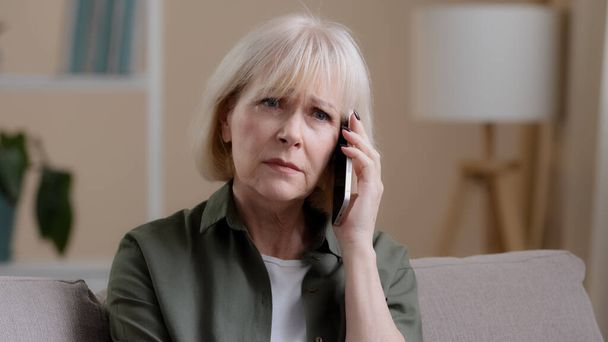 Caucasian old woman mature middle-aged 50s offended lady aged businesswoman sitting on sofa at home listening to voice on phone talking smartphone receiving bad news sadness worry upset losing failure - Foto, Imagen