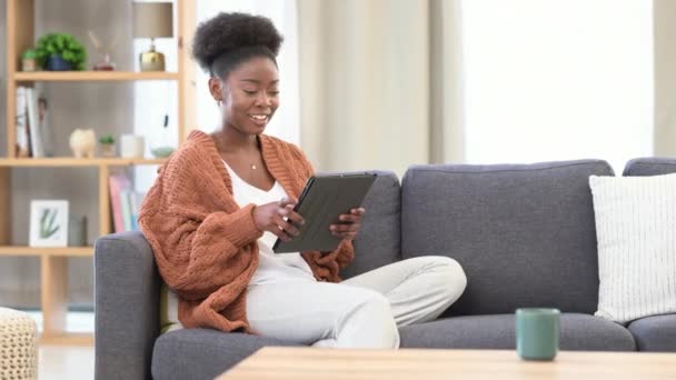 Woman laughing and browsing social media online on a digital tablet while relaxing on the couch at home. One young black female smiling while searching the internet and texting friends on the web. - Filmagem, Vídeo