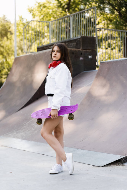 Child girl with penny board and headphones posing on skate sport ramp. Sports equipment for kids. Active teenager with penny board on skate park playground - 写真・画像