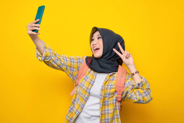 Smiling young Asian Muslim woman student in Plaid shirt and backpack using mobile phone take selfie make peace sign isolated on yellow background. Education school university college concept - Foto, imagen