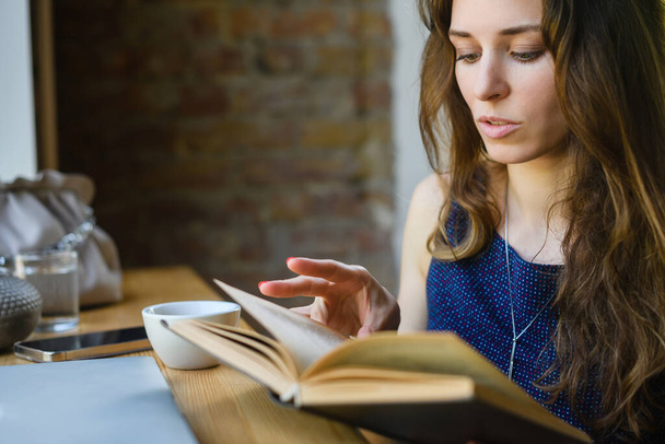Attractive young girl reading a book while having breakfast in a coffee shop. Close-up. - Foto, Imagen