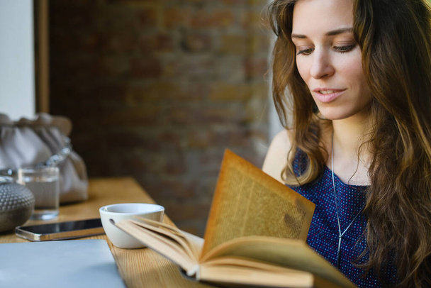 Attractive young girl reading a book while having breakfast in a coffee shop. Close-up. - Foto, Imagen