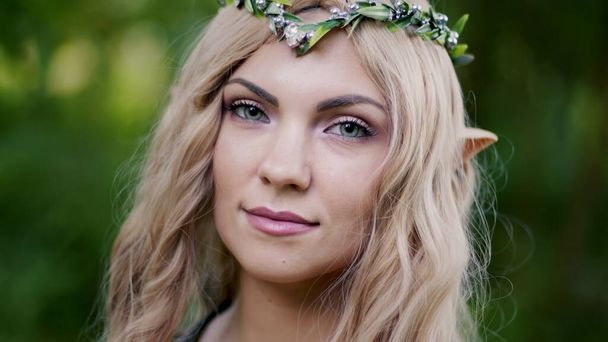 Portrait of young woman in cosplay elf clothes with make-up on green background. Fantastic look, long blonde hair, forest crown. Halloween concept. High quality photo - Фото, изображение