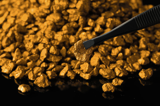 Tweezers with gold nugget above pile on black background, closeup - Foto, Imagen
