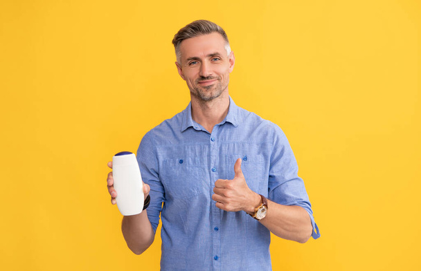 volwassen man met grizzled haar hold shampoo fles show duim op gele achtergrond, reclame. - Foto, afbeelding