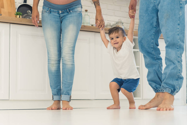 Mutfakta çıplak ayakla yürüyen üç kişilik bir aile. Küçük çocuk zıplamak için anne ve babasının arasında durup ellerini tutuyor. Yüksek kalite fotoğraf - Fotoğraf, Görsel