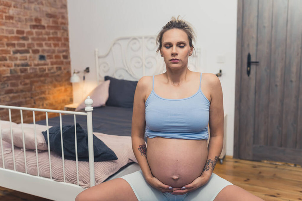 Young pregnant caucasian mother touching her belly and exercising on a fitness ball. Bedroom interior. High quality photo - Foto, afbeelding