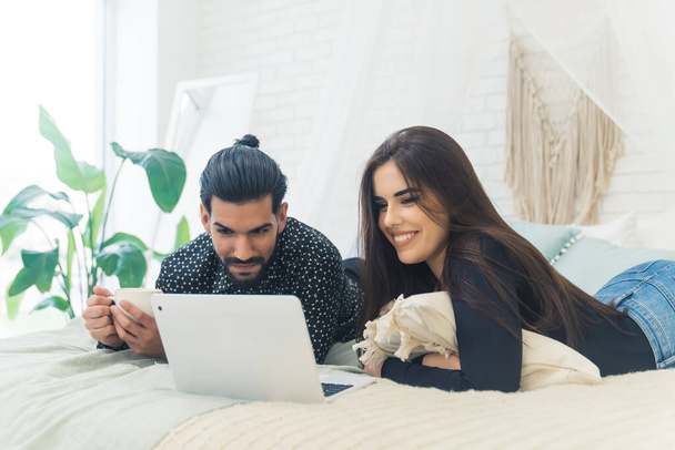 Adorable happy heterosexual couple watching an online available series and laughing while lying down on their bed in a modernly decorated bedroom. Free time activities. . High quality photo - Photo, image