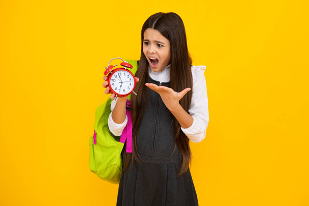 Back to school. Teenage school girl with bag hold clock alarm, time to learn. Schoolgirl on isolated yellow background. Angry teenager girl, upset and unhappy negative emotion - Foto, afbeelding