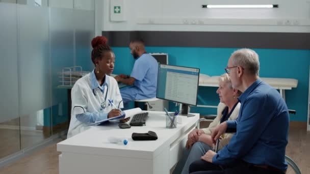 African american medic talking to elderly woman with disability at chekcup visit appointment. Senior patient with impairment in wheelchair and husband receiving medical disease treatment. - Materiał filmowy, wideo