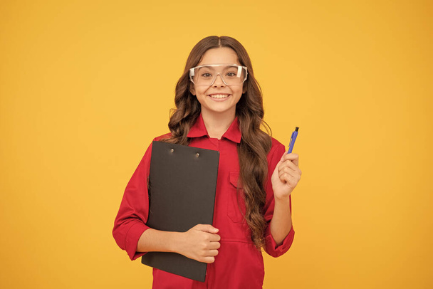 gotowy z wynikami. Szczęśliwego dzieciństwa. Nastolatka nosi okulary. Dzieciak robi notatki w folderze na żółtym tle. nauka dzieci w szkole. pracować z dokumentami. uśmiechnięty dzieciak w okularach ochronnych. - Zdjęcie, obraz