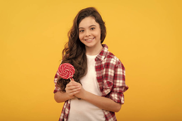 fröhliche Teenie-Mädchen mit langen lockigen Haaren im karierten Hemd halten Lutscher Karamellbonbons auf gelbem Hintergrund, Karamellladen. - Foto, Bild