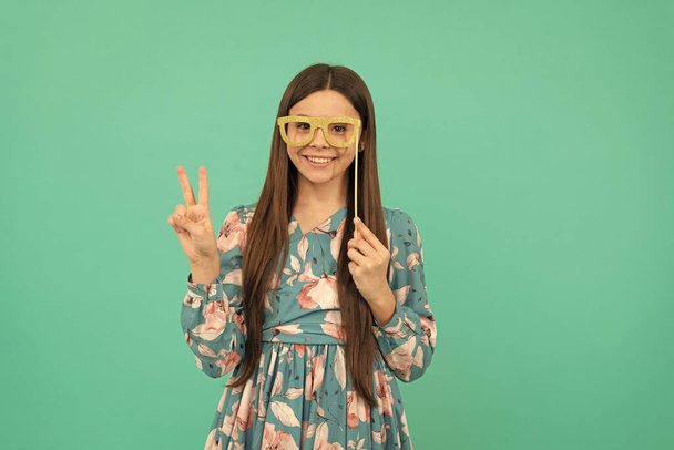 Feliz niña sostiene gafas de utilería que muestran victoria mano v-signo fondo azul, fresco. - Foto, Imagen