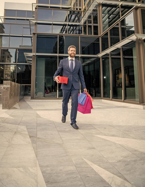 vrolijke volwassen zakenman in formele kleding wandeling buiten met boodschappentassen voor vakantie, volledige lengte, verjaardag. - Foto, afbeelding