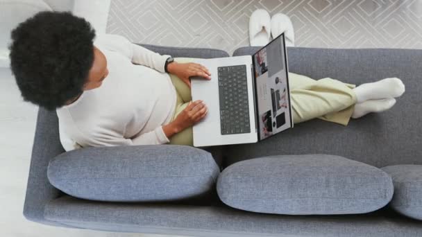Online shopping on laptop in home living room or browsing variety of options in lockdown. Above view of relaxed woman with afro using technology to blog, trade or study distance learning course. - Video