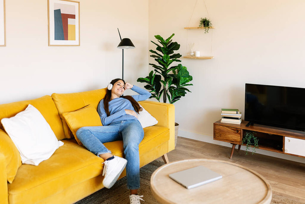 Relaxed smiling young adult woman sitting on sofa while listening to music at home - 写真・画像