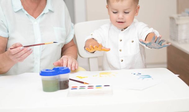 People. Caucasoid woman and a little boy in light clothes draw with paints at a white table in a home interior. Grandmother and grandson. Soft focus. - Photo, Image