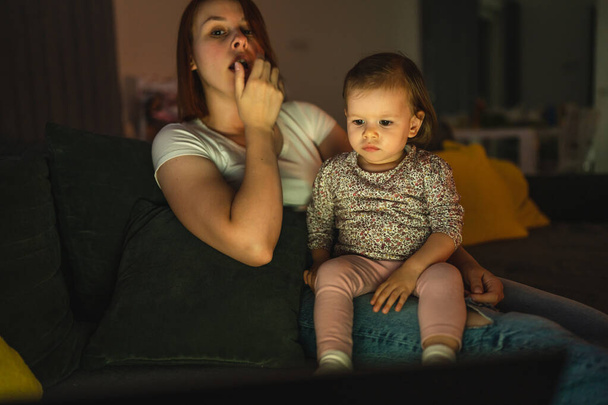 Маленький кавказский ребенок маленькая девочка малышка мать и дочь сидит в темной комнате ночью дома на диване кровать смотреть мультфильмы или фильмы на ноутбуке реальные люди сонные семейное пространство копирования - Фото, изображение