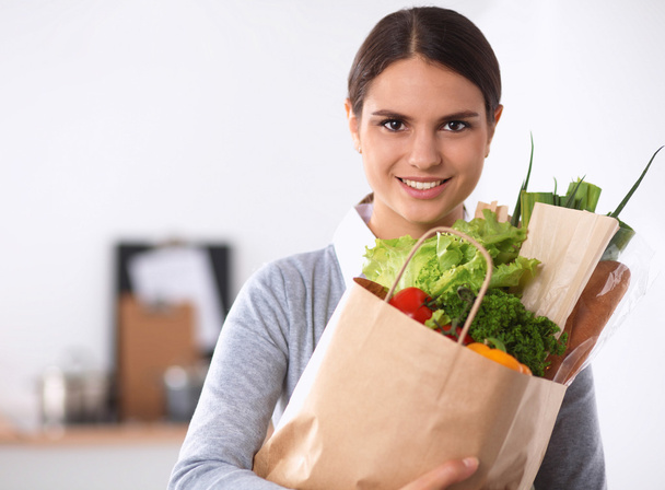 野菜と食料品の買い物袋を持っている若い女性キッチンに立つ. - 写真・画像