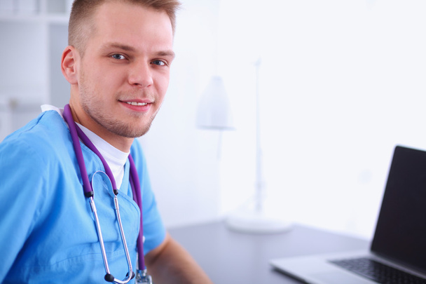 Portrait of a smiling male doctor with laptop sitting at desk in medical office - Photo, Image