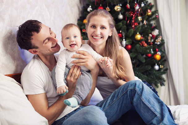 Familia joven con un bebé divirtiéndose en la cama en casa con árbol de Navidad en el fondo
 - Foto, Imagen